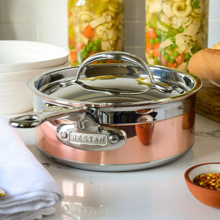 A sleek CopperBond Copper Induction Saucepan with a lid elegantly graces the counter, flaunting its stainless steel design and copper core finish. Nearby, jars of pasta, vegetables, a white towel, a bowl, and chili flakes sit ready. Ideal for all induction cookware needs.