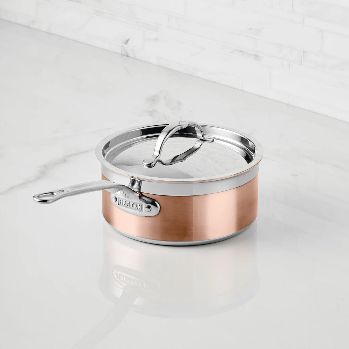 A CopperBond Copper Induction Saucepan with a stainless steel lid sits on a white marble countertop, set against a white subway tile backsplash.