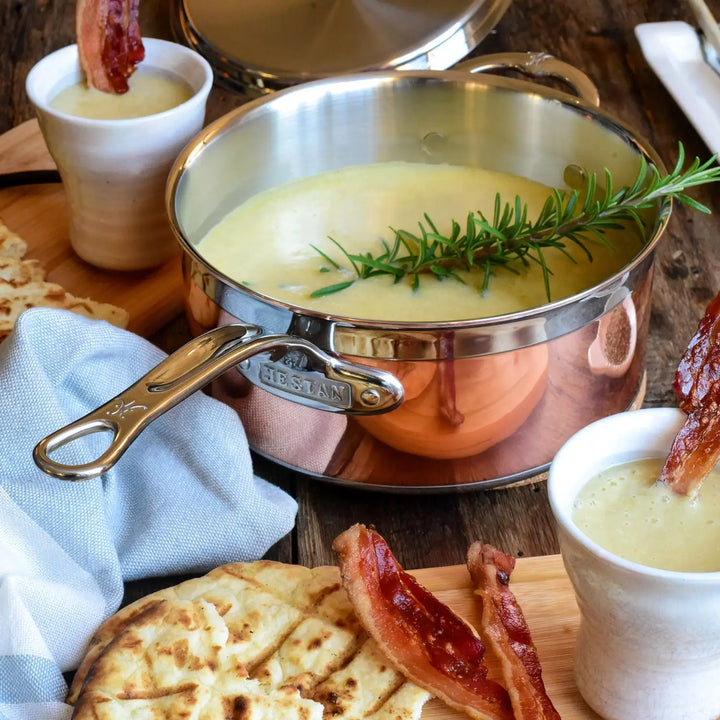 In a CopperBond Copper Induction Saucepan, creamy soup with rosemary sits on a wooden surface. Two bacon-adorned cups of soup are nearby, along with flatbreads and extra bacon. A lid and cloth in the background emphasize the sophistication of this induction cookware.