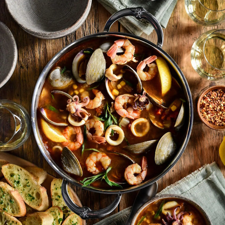 A CopperBond Copper Induction Stock Pot, 6-Quart, sits on a wooden table brimming with seafood stew—shrimp, clams, and squid rings mingling with herbs and lemon wedges. Surrounding this 5-layer stainless steel marvel are slices of garlic bread, filled wine glasses, and a small bowl of red pepper flakes.