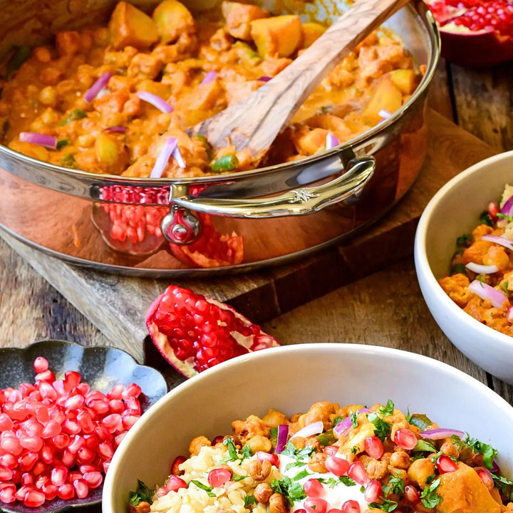 A curry in a CopperBond Copper Induction Sauté with Lid, 3.5-Quart, sits on a wood tray with vegetable chunks. Nearby are pomegranate halves and seeds in a dish. In the foreground, two bowls are filled with rice, chickpeas, herbs, onion slivers, and pomegranate seeds.