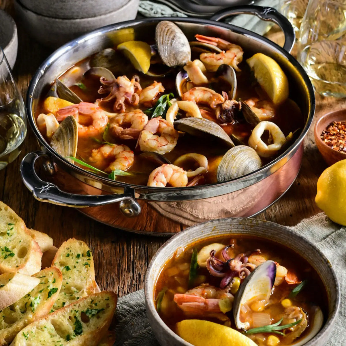 A pot of seafood stew with shrimp, clams, and squid sits beside a bowl of stew, lemon wedges, toasted bread, and white wine—served in a 3.5-quart CopperBond Copper Induction Braiser with Lid.