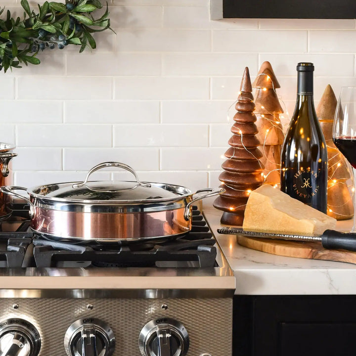 A kitchen counter features a stainless steel pot on a stove with knobs, surrounded by a large cheese wedge, the CopperBond 3.5-Quart Copper Induction Sauteuse with Lid, a grater, and red wine next to a full glass. Decorative wooden trees with string lights add charm—a perfect culinary setting.