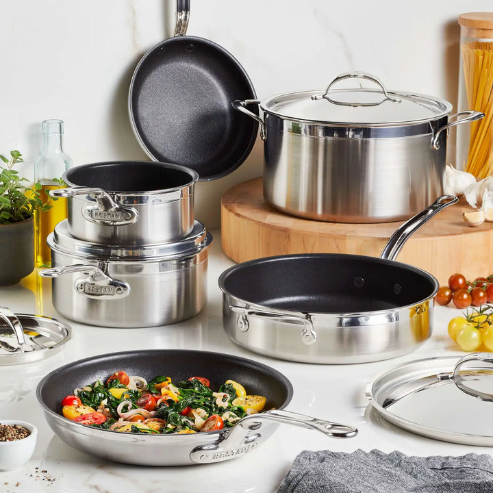 A kitchen scene displays ProBonds Professional Clad Stainless Steel TITUM® Nonstick Ultimate Cookware Set, featuring skillets and a large saucepan, some with lids. One pan holds cooked vegetables while olive oil, spices, and uncooked pasta sit on the marble countertop.