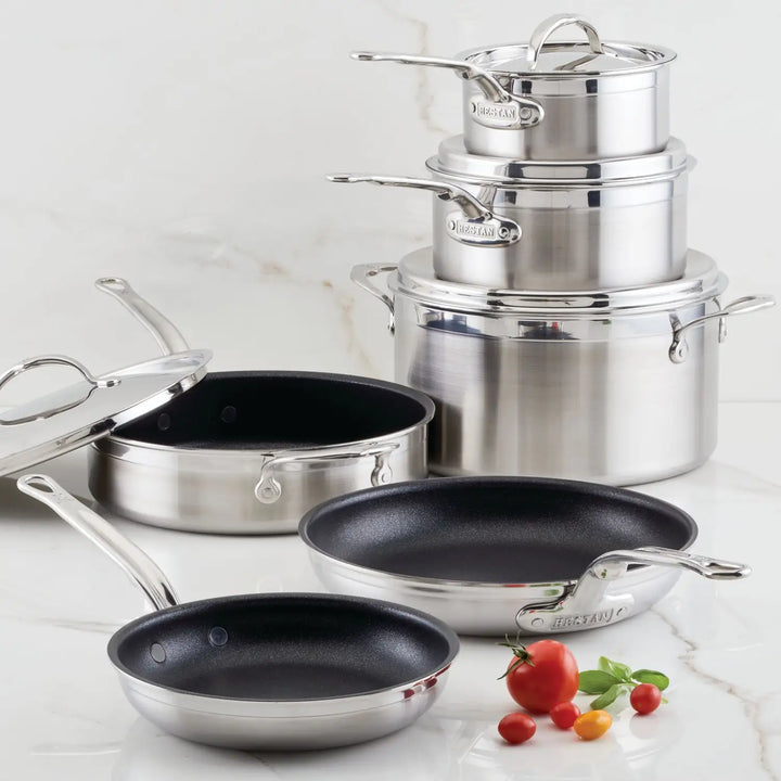 A ProBond Professional Clad Stainless Steel TITUM® Nonstick Ultimate Cookware Set, 10-piece, featuring saucepans with lids, a stockpot, and nonstick frying pans with black interiors is displayed on a white marble surface with two cherry tomatoes and a small bell pepper in the foreground.