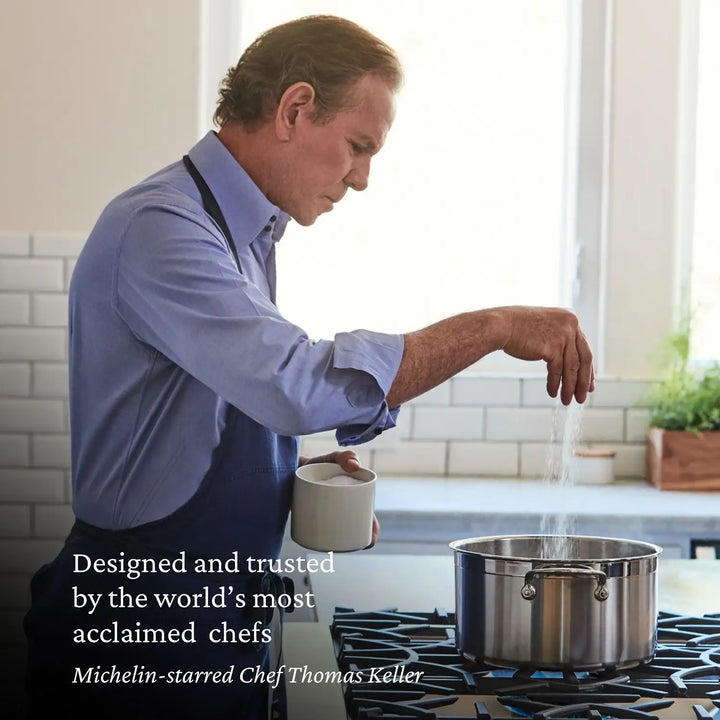 A Michelin-starred chef, wearing a blue shirt and apron, skillfully adds salt to a pot on the stove while holding a white mug. The bright kitchen shines with the ProBond Professional Clad Stainless Steel TITUM® Nonstick Ultimate Cookware Set, enhancing the culinary artistry on display.