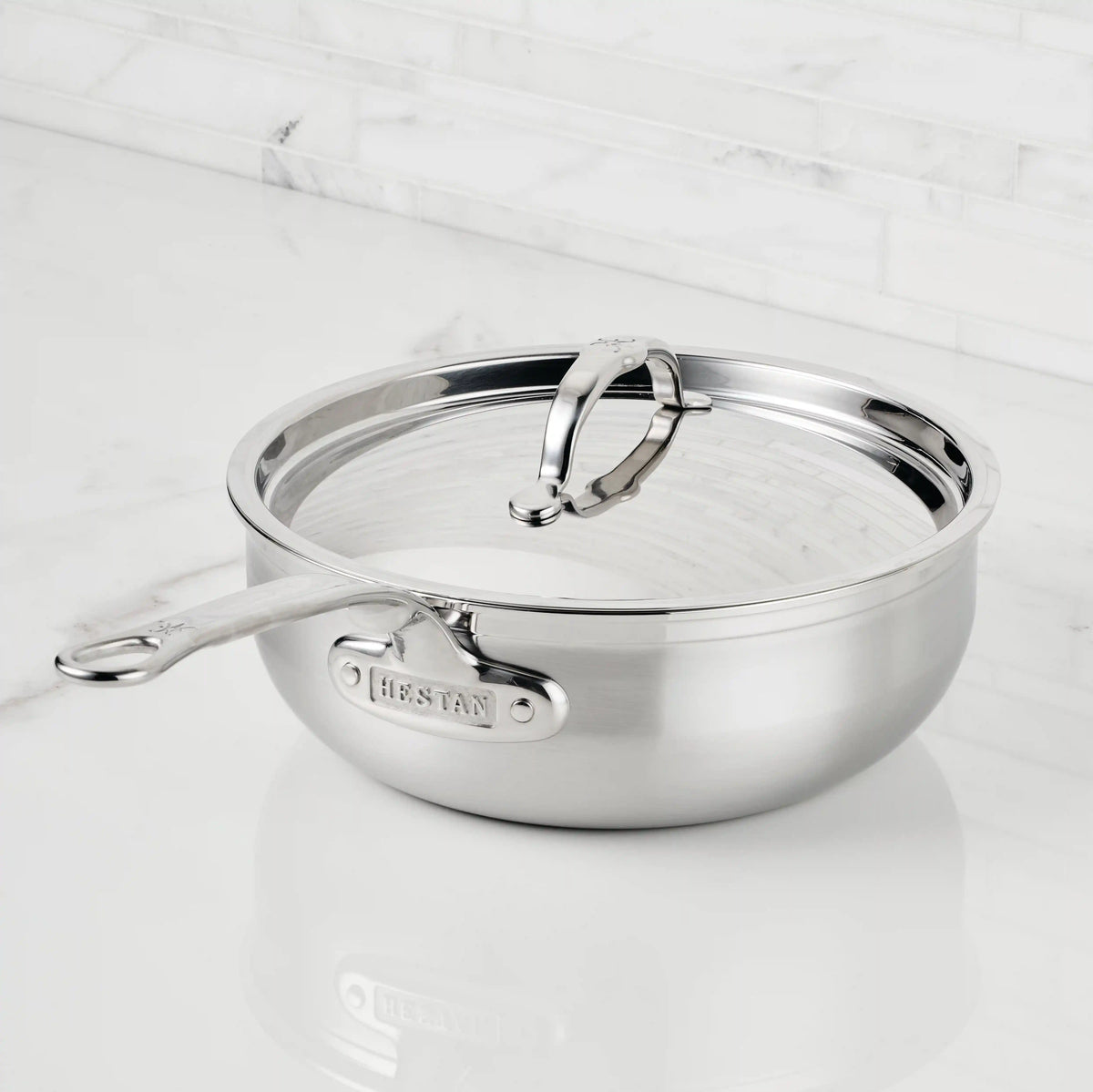 A ProBond® Forged™ Professional Clad Stainless Steel Essential Pan with a long handle and matching lid sits on a white marble countertop, showcasing its premium design and visible brand engraving.