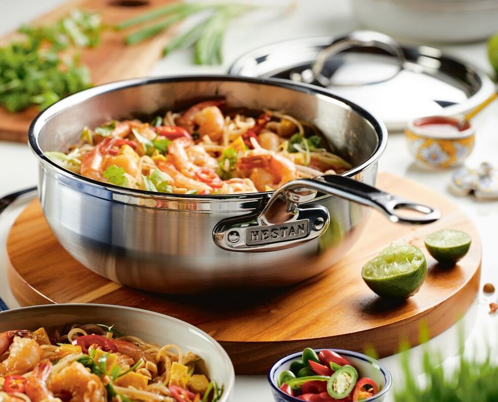 A ProBond Professional Clad Stainless Steel Induction pot from a 4pc Cookware Set holds a stir-fry of shrimp, vegetables, and noodles on wood. Nearby are sliced limes, chili peppers, and greens.