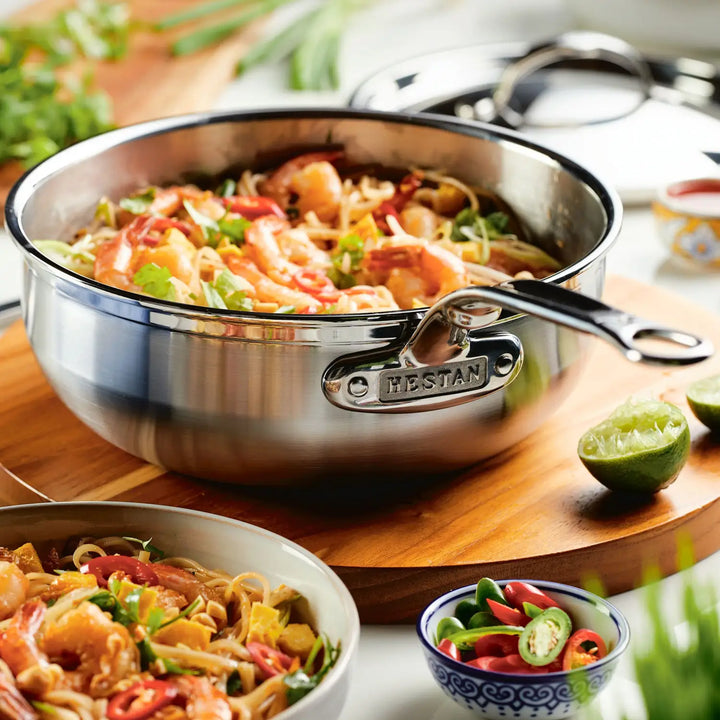 A ProBond Professional Clad Stainless Steel pot with tri-ply aluminum core holds shrimp, noodles, and veggies. Beside it, a ProBond Essential Pan contains a similar dish; sliced chilies are in a nearby dish with half a lime visible in the background.