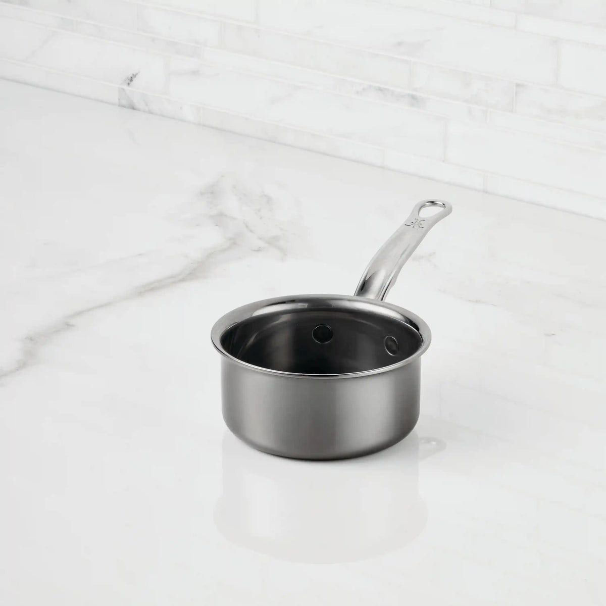 A NanoBond Titanium Butter Warmer (.75 quarts) with a long handle sits on a white marble countertop against a tiled backsplash, making it an elegant and stylish choice for warming butter.