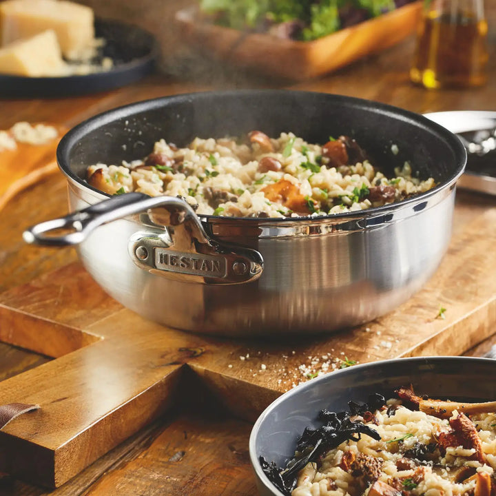 A ProBond Clad Stainless Steel TITUM® Nonstick Essential Pan, 3.5-Quart, filled with steaming mushroom risotto and garnished with herbs, sits on a wooden cutting board. In the foreground is a small bowl with a serving of the risotto.