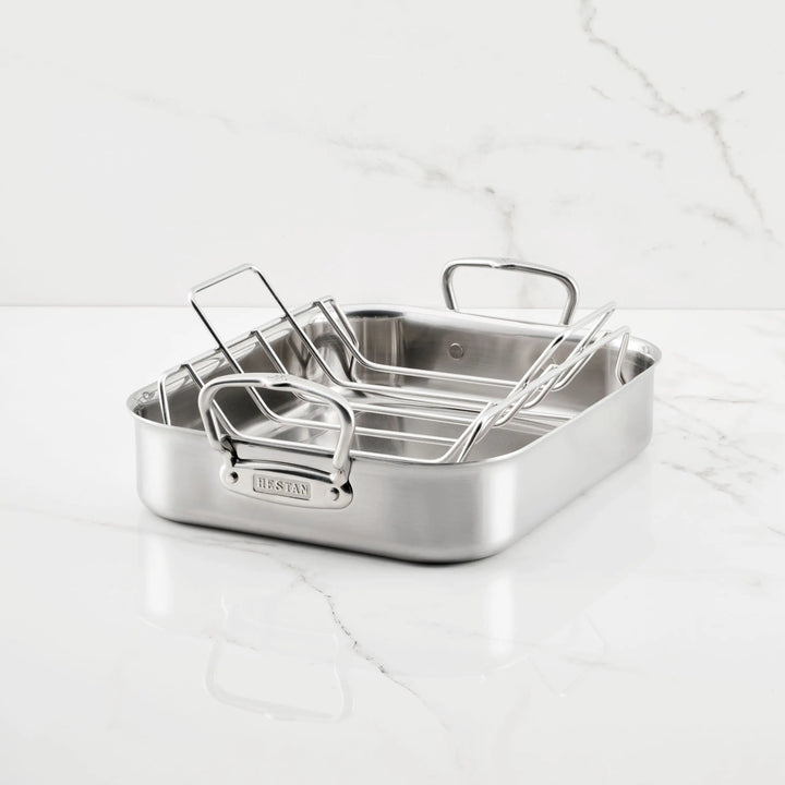 The Hestan Provisions Brushed Clad Stainless Steel Roaster with Rack, featuring two upright handles, sits on a white marble countertop with gray veining.