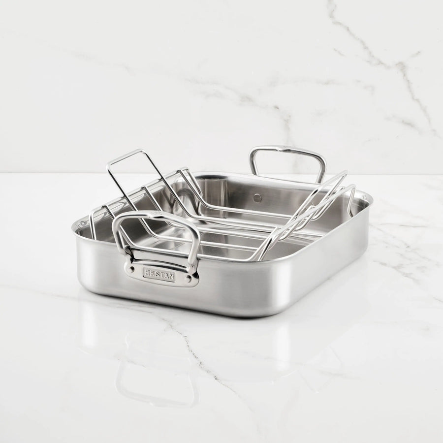 The Hestan Provisions Brushed Clad Stainless Steel Roaster with Rack, featuring two upright handles, sits on a white marble countertop with gray veining.