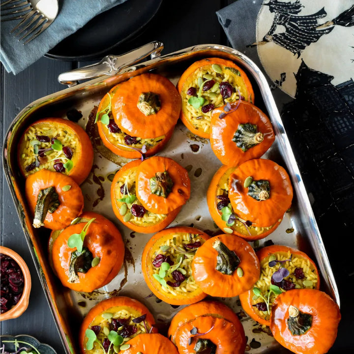 A Hestan Provisions 16.5-inch Classic Clad Roaster with Rack is filled with small pumpkins, each stuffed with grains and topped with herbs and cranberries. A printed cloth napkin and a bowl of cranberries are placed nearby.