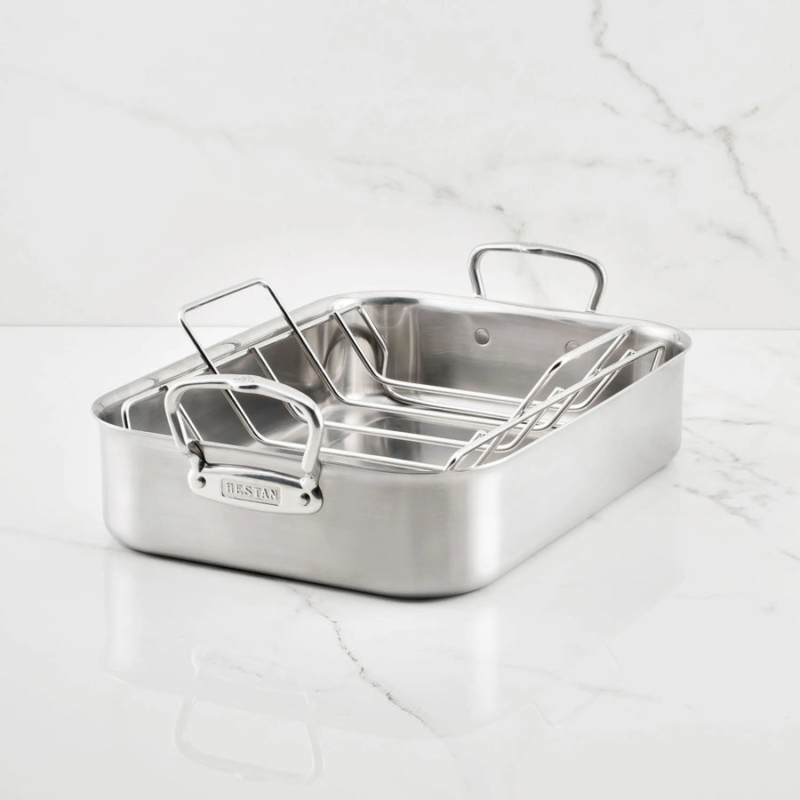 A Hestan Provisions Brushed Clad Stainless Steel Roaster with handles and a removable rack rests on a white marble surface. The rectangular pan showcases a sleek brushed finish.