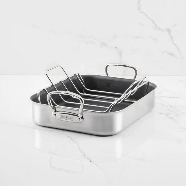 The Hestan Provisions Nonstick Clad Stainless Steel Roaster with Rack, featuring two handles and an elevated rack, rests on a white marble surface with a light gray veined background.