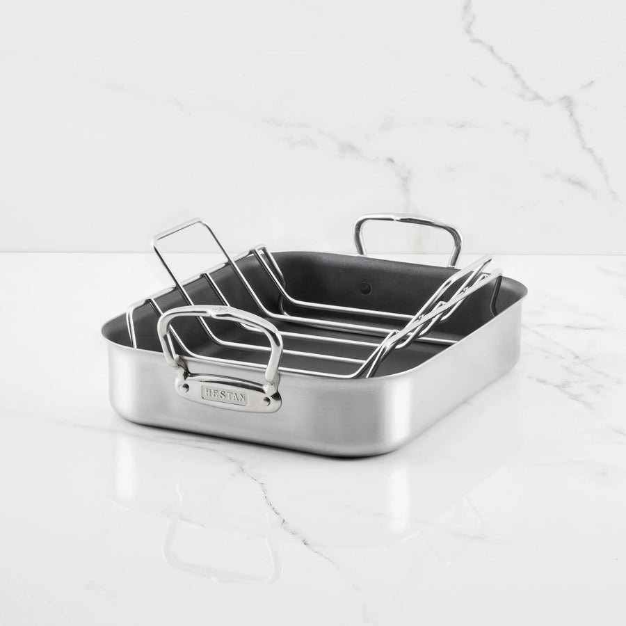 The Hestan Provisions Nonstick Clad Stainless Steel Roaster with Rack, featuring two handles and an elevated rack, rests on a white marble surface with a light gray veined background.