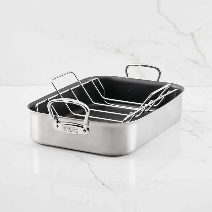 The Hestan Provisions Nonstick Clad Stainless Steel Roasters with Rack, featuring two large handles, is displayed on a white marble surface with gray veining—a must-have addition to your stainless steel cookware collection.