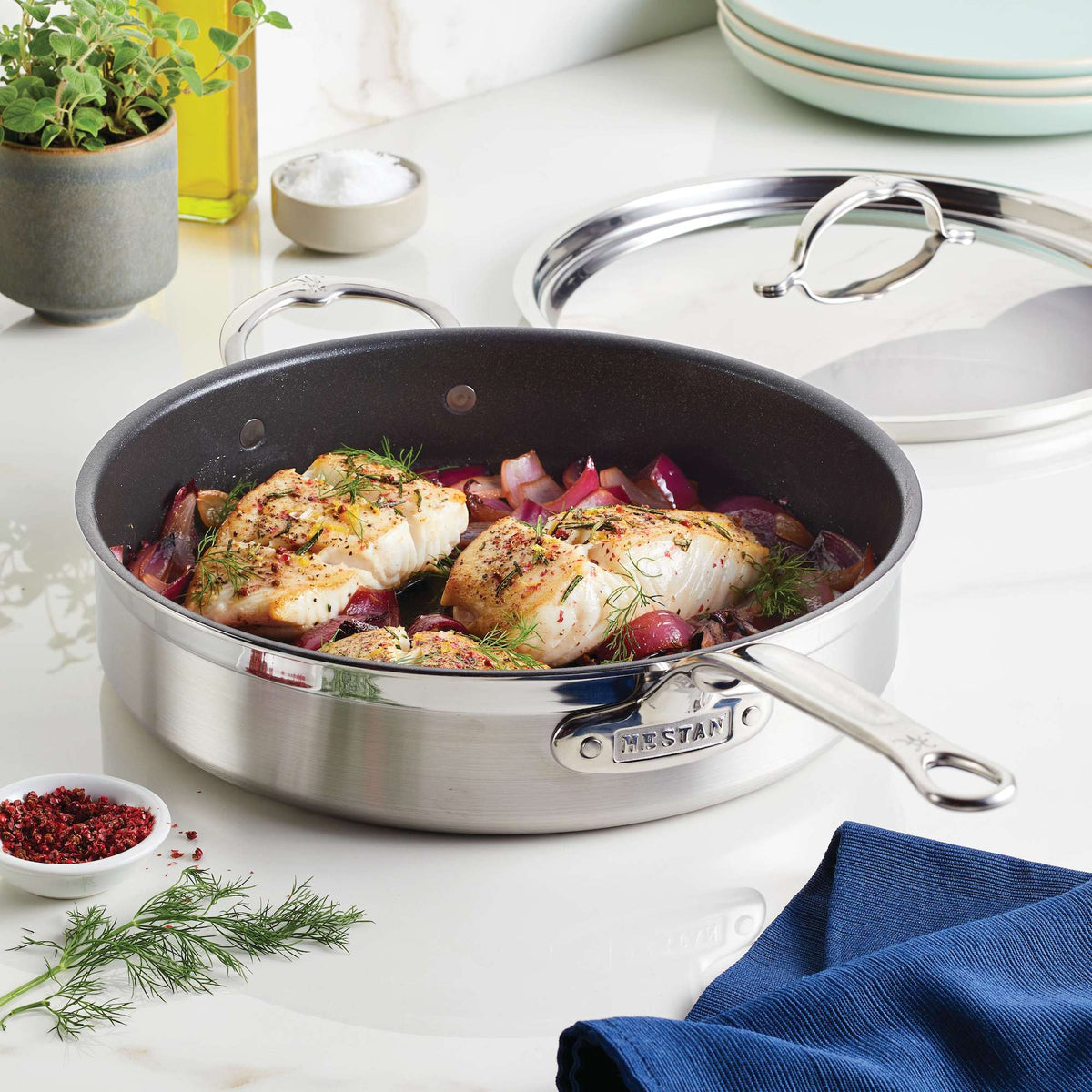 A ProBond 5-Quart Professional Clad Stainless Steel TITUM® Nonstick Sauté Pan containing cooked chicken breasts with herbs and red onions rests on a countertop. Nearby, its lid sits next to a plant pot, an oil bottle, dinnerware, and a blue cloth that adds color.