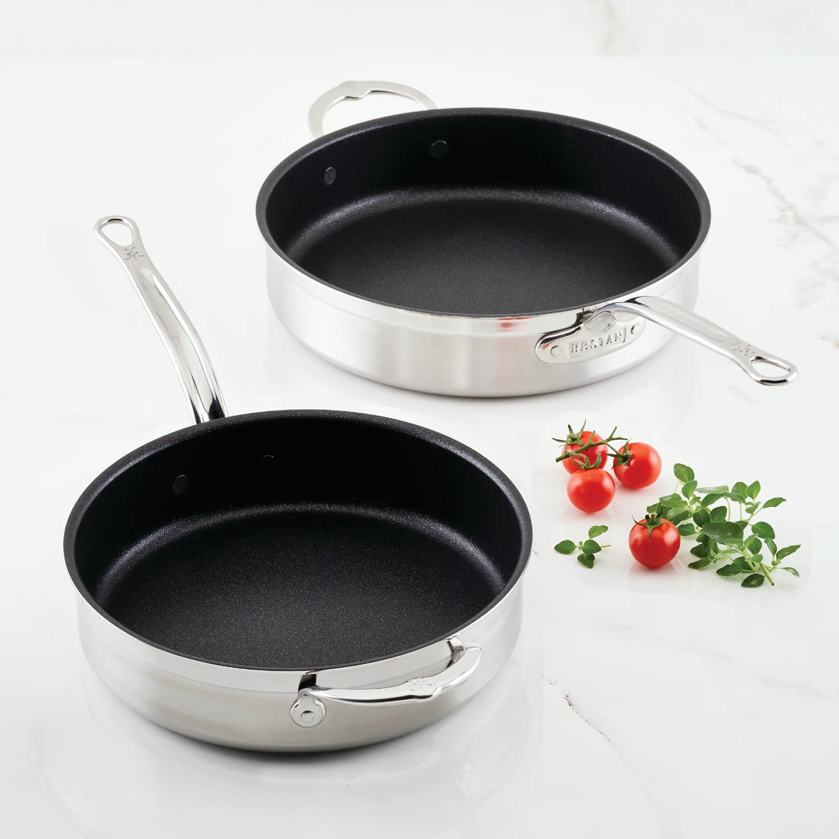 Two ProBond sauté pans with black TITUM® non-stick interiors rest on a white marble surface. Crafted for precision, one pan is slightly smaller. Next to them are three red cherry tomatoes and a small sprig of green herbs.
