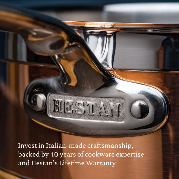 A close-up of a shiny metallic saucepan with a handle attached by rivets displays the embossed HESTAN text. This CopperBond Copper Induction Sauté with Lid, 3.5-Quart, embodies Italian craftsmanship, 40 years of expertise, induction compatibility, and comes with a lifetime warranty.