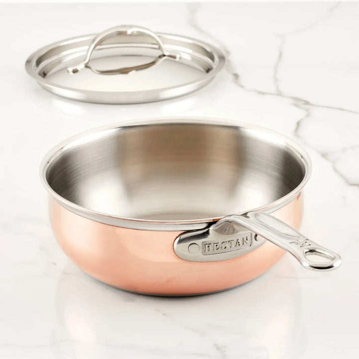 A CopperBond Copper Induction Saucier Pan, 2-Quart with a stainless steel interior sits on marble. It has a metal handle and comes with a matching lid elegantly positioned behind it.