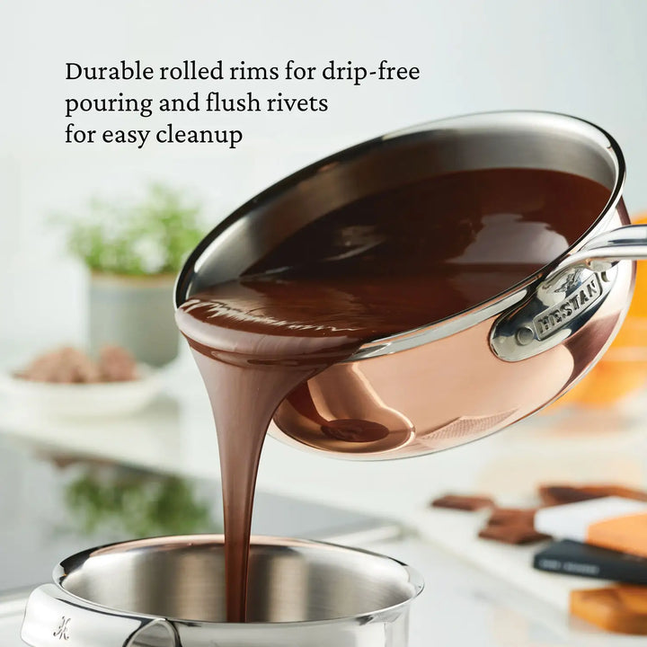 A CopperBond Copper Induction Saucier Pan, 2-Quart elegantly pours chocolate sauce, featuring durable rolled rims, flush rivets, and a sturdy steel handle with the brand name. The blurred kitchen background adds depth, highlighting its versatile induction compatibility.