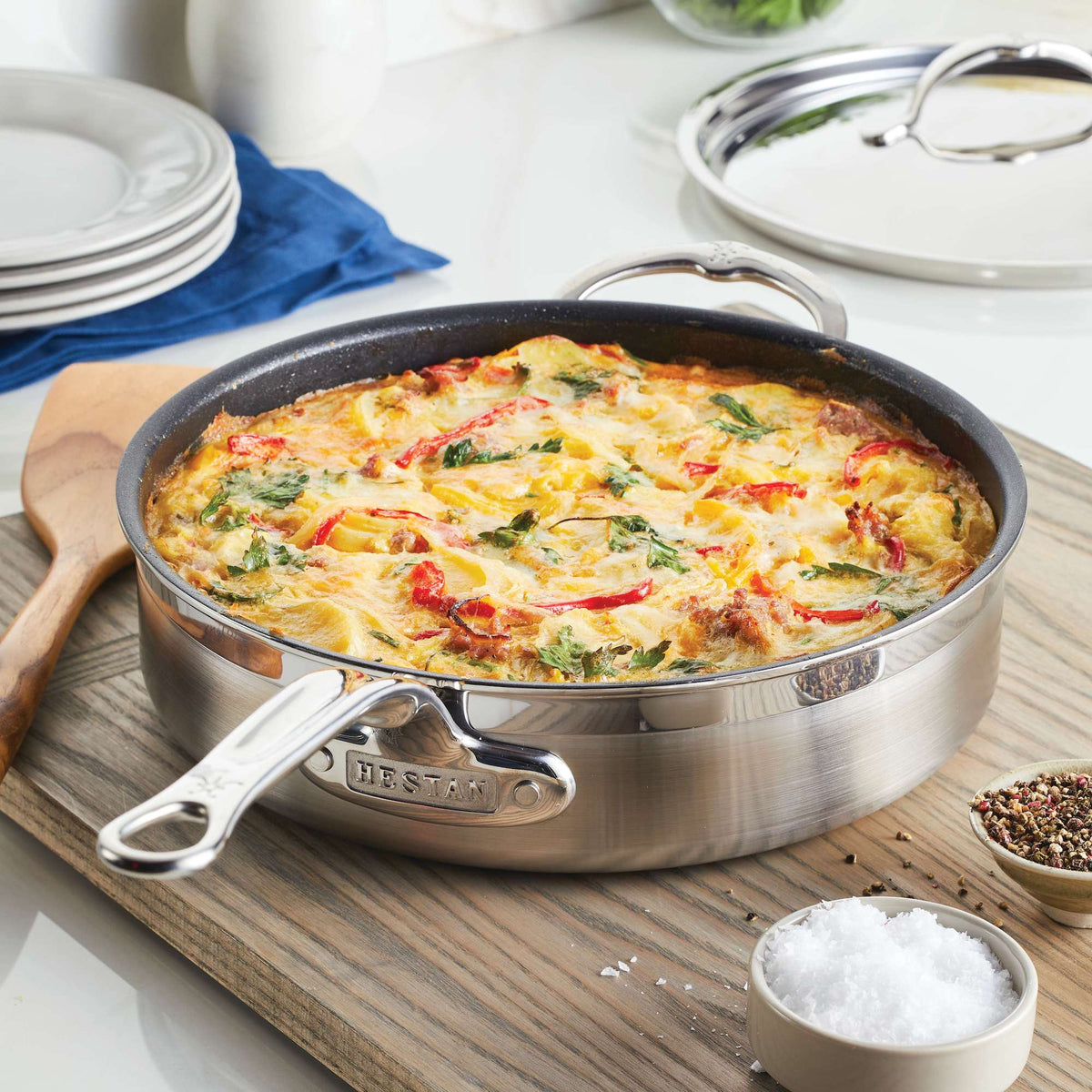 A colorful frittata in a ProBonds 3.5-quart TITUM® Nonstick Sauté Pan rests on a wooden surface, featuring vibrant vegetables and melted cheese. Nearby, find a wooden spatula, salt container, pepper grinder, and stacked plates with a blue napkin.