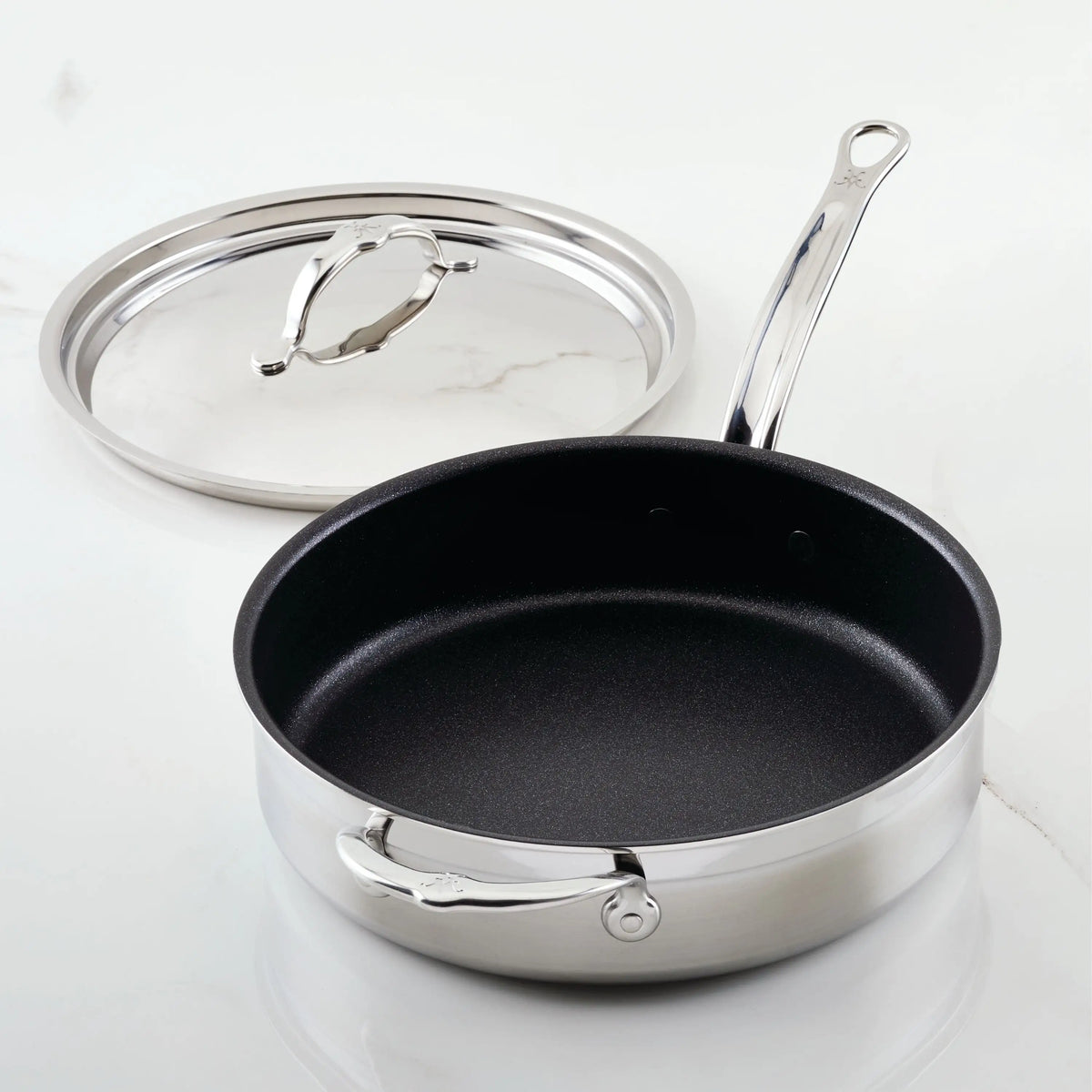 The sleek 3.5-Quart ProBond Professional Clad Stainless Steel TITUM® Nonstick Sauté Pan with Lid rests on a light surface, boasting a long handle and smaller helper handle. Its matching lid is elegantly placed beside this Hestan masterpiece.
