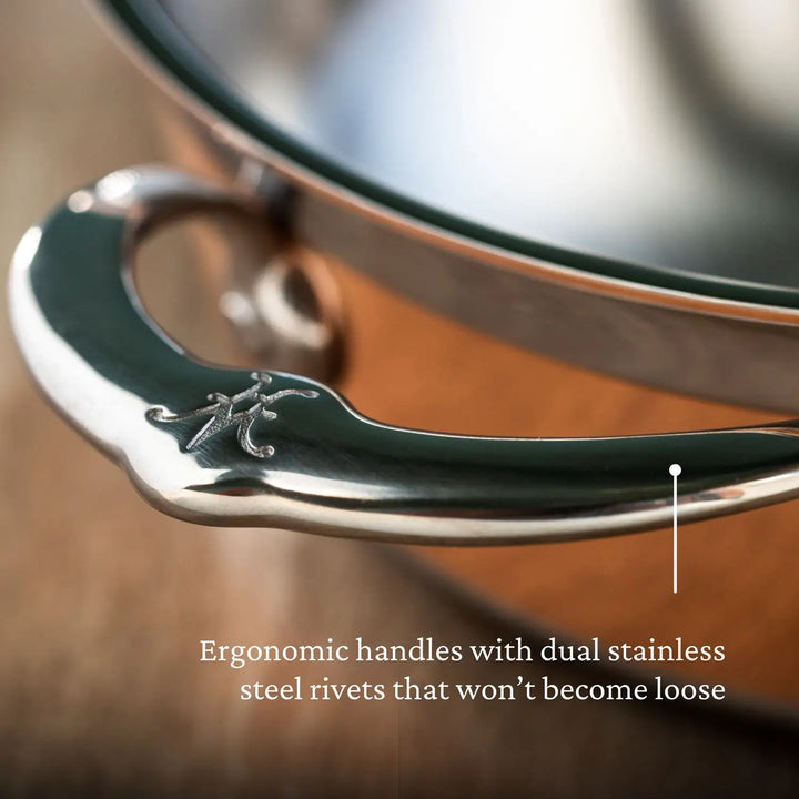 Close-up of a CopperBond Dutch Oven handle. The metallic design, ideal for the Copper Induction Dutch Oven, 5-Quart, features an engraved logo with intertwined letters. Text reads: Ergonomic handles with dual stainless steel rivets that won’t become loose. Background is blurred.