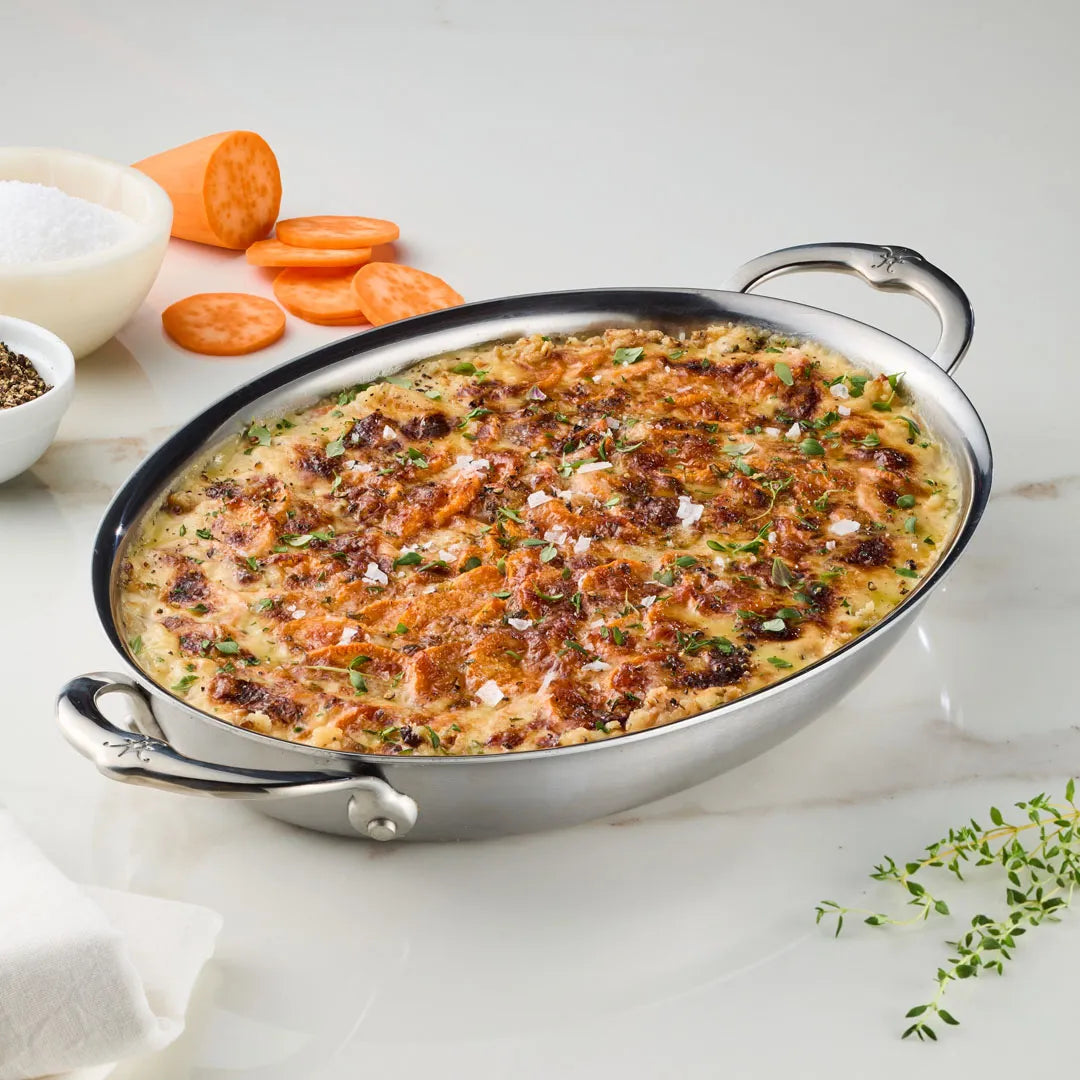 A Hestan Provisions 12.5-Inch x 9-Inch Oval Au Gratin dish holds golden sweet potato and herb gratin on a white countertop, with sliced sweet potatoes, salt, pepper bowls, and fresh thyme nearby.