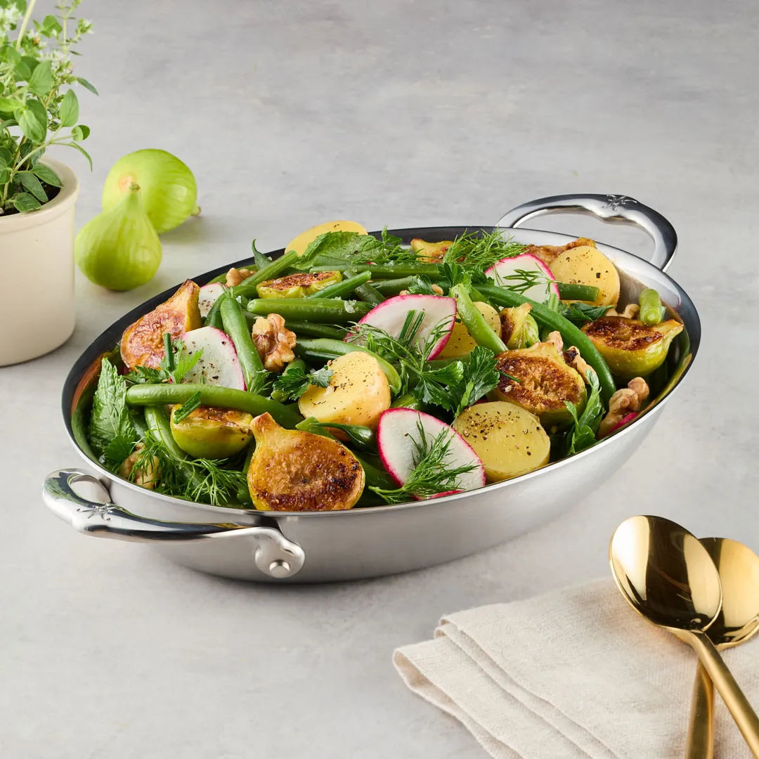 A Hestan Provisions 12.5-Inch x 9-Inch Oval Au Gratin filled with green beans, potatoes, radishes, figs, walnuts, and fresh herbs sits on a light surface beside a potted plant, folded napkin, and gold-colored utensils.