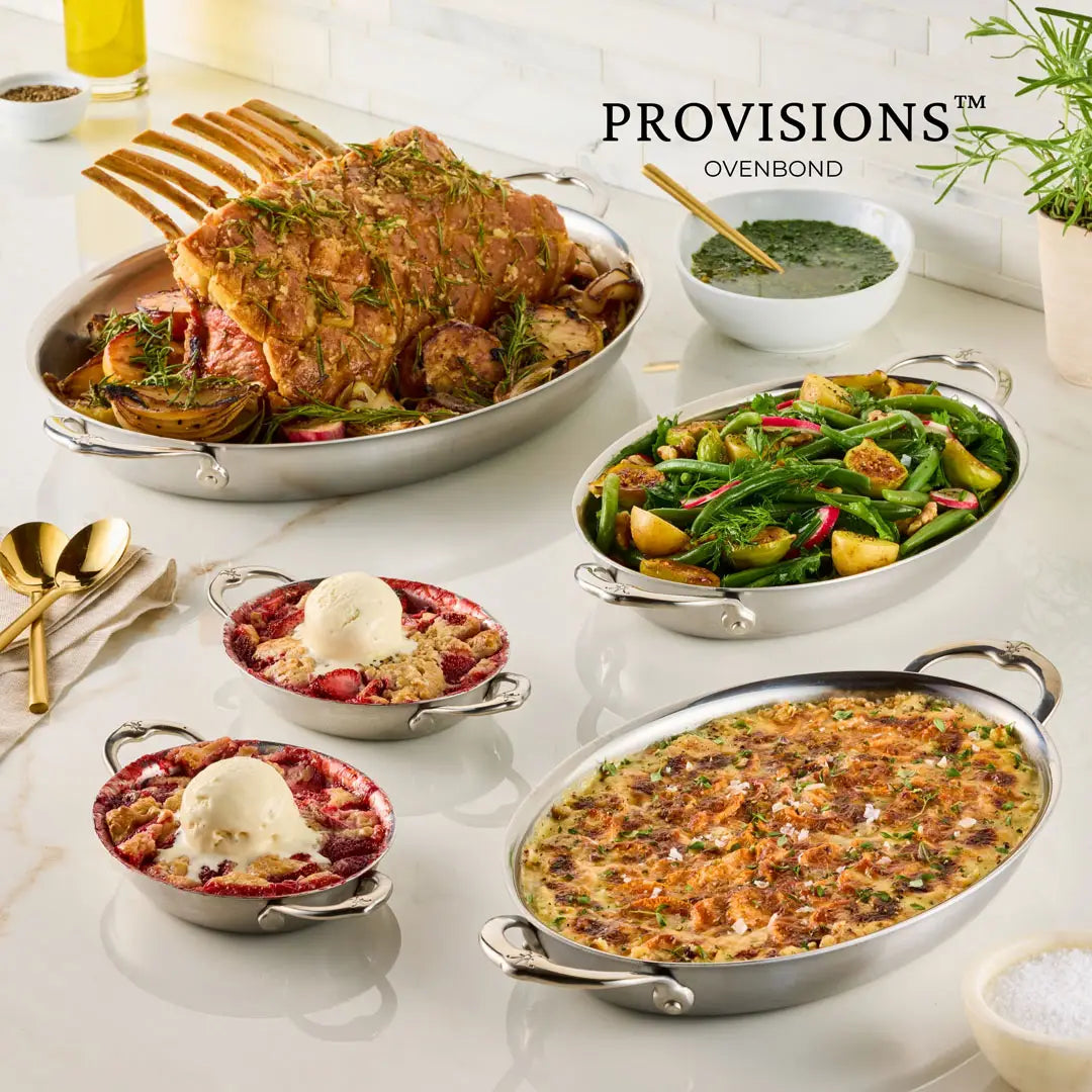 A table set with Hestan Provisions chef-designed bakeware displays roast meat with herbs, mixed roasted vegetables, a green vegetable dish, and a casserole in the 15-Inch x 9-Inch Oval Au Gratin, plus two berry desserts with ice cream. Gold utensils accompany the spread.