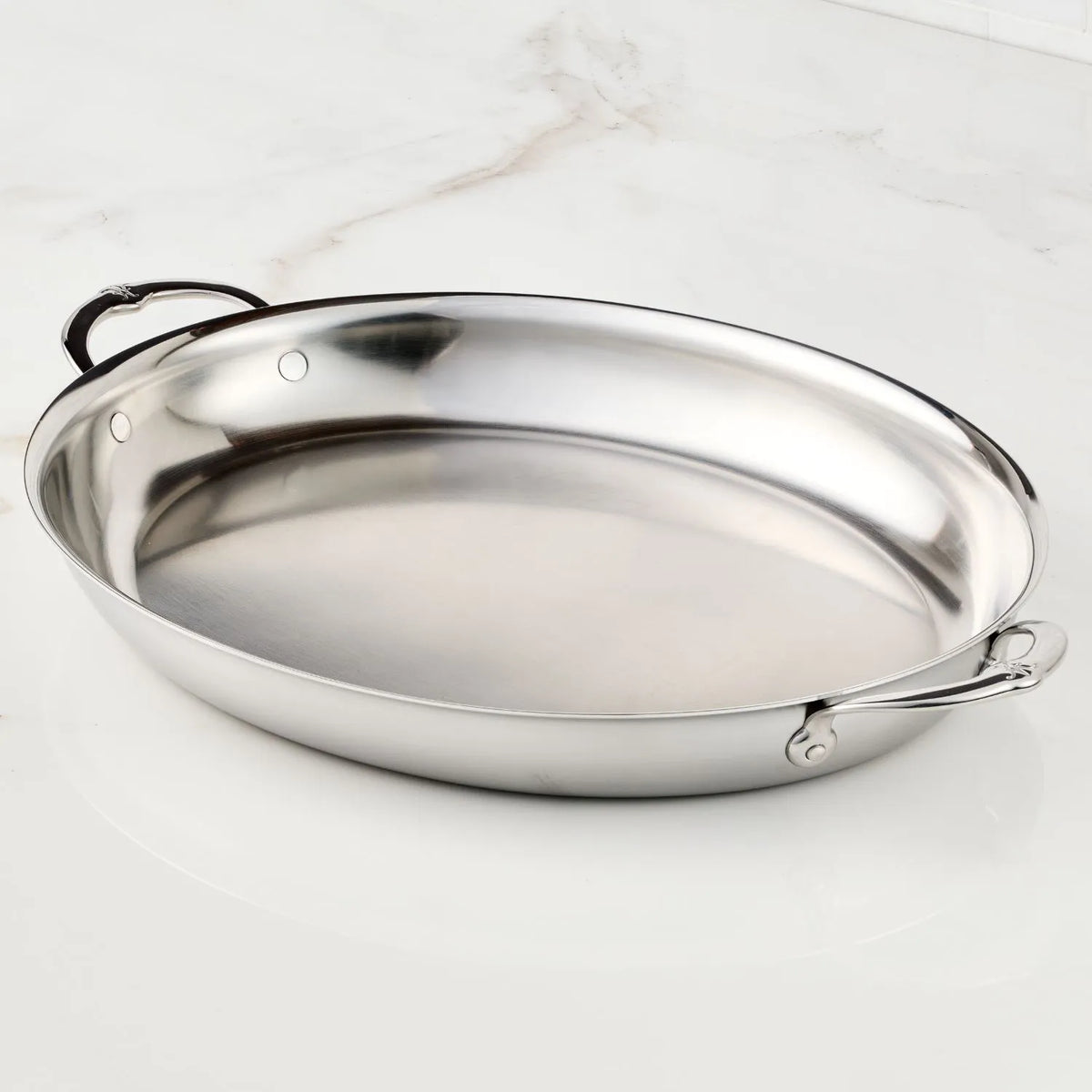 The Hestan Provisions 17.5-Inch x 14-Inch Clad Oval Roaster, made of stainless steel with two handles and a shiny, reflective finish, rests empty on a white marble surface, highlighting its quality craftsmanship.