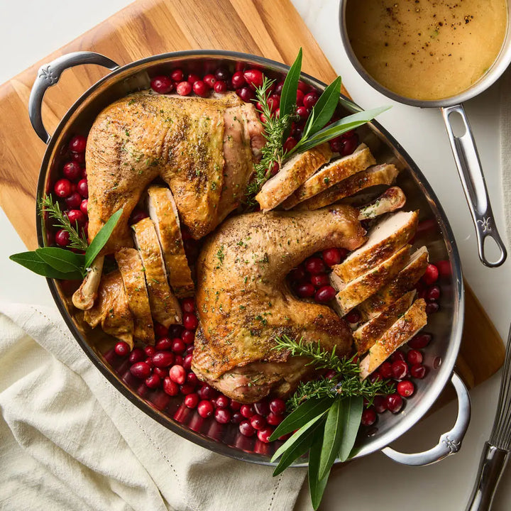 A roasted chicken with sliced breasts and whole legs is arranged on a bed of cranberries in a Hestan Provisions Large Clad Oval Roaster. Fresh herbs adorn the top. Nearby, a saucepan with gravy sits on a wooden board, demonstrating the finesse of Michelin-starred Chef Thomas Keller.