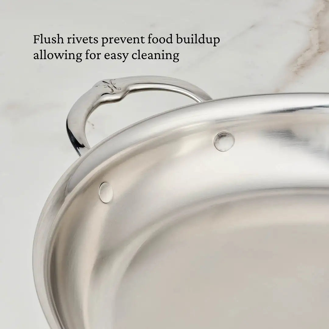A close-up of the Hestan Provisions 17.5-Inch x 14-Inch Clad Oval Roaster shows its stainless steel interior and handle with flush rivets. Text on image: Flush rivets prevent food buildup, allowing for easy cleaning.