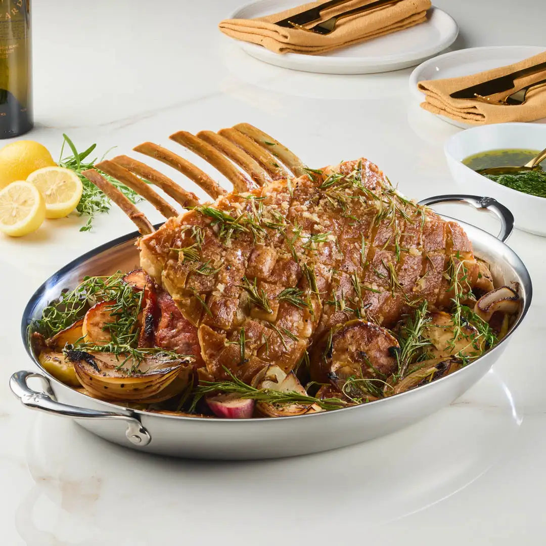 A cooked rack of lamb with herbs sits in a Hestan Provisions 17.5-Inch x 14-Inch Clad Oval Roaster, surrounded by roasted onions and potatoes, with lemon halves, plates, napkins, utensils, and green sauce visible in the background.