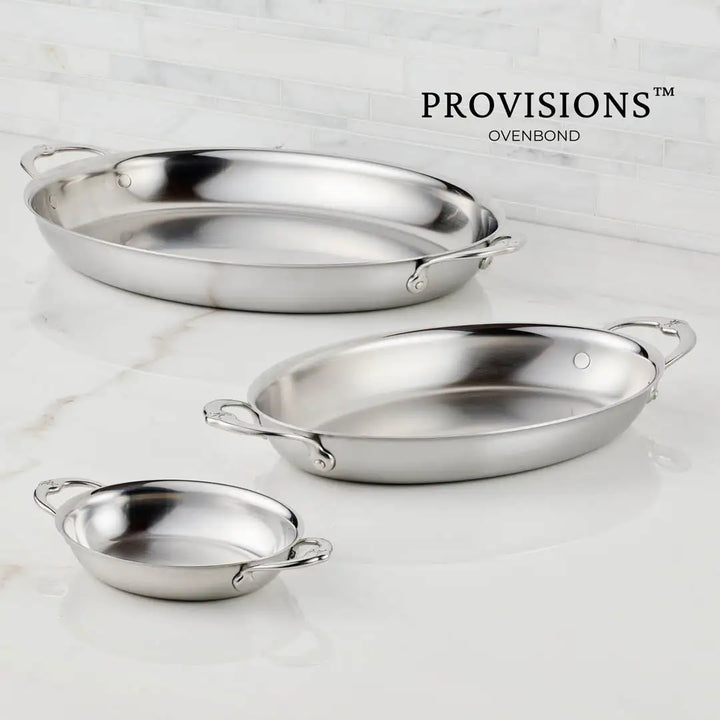 Three Hestan Provisions Large Clad Oval Roasters are elegantly displayed on a white marble surface. Designed for Michelin-starred chefs, these stainless steel pans vary in size: the largest at the back, medium in the middle, and smallest upfront. Sleek handles enhance their sophistication.