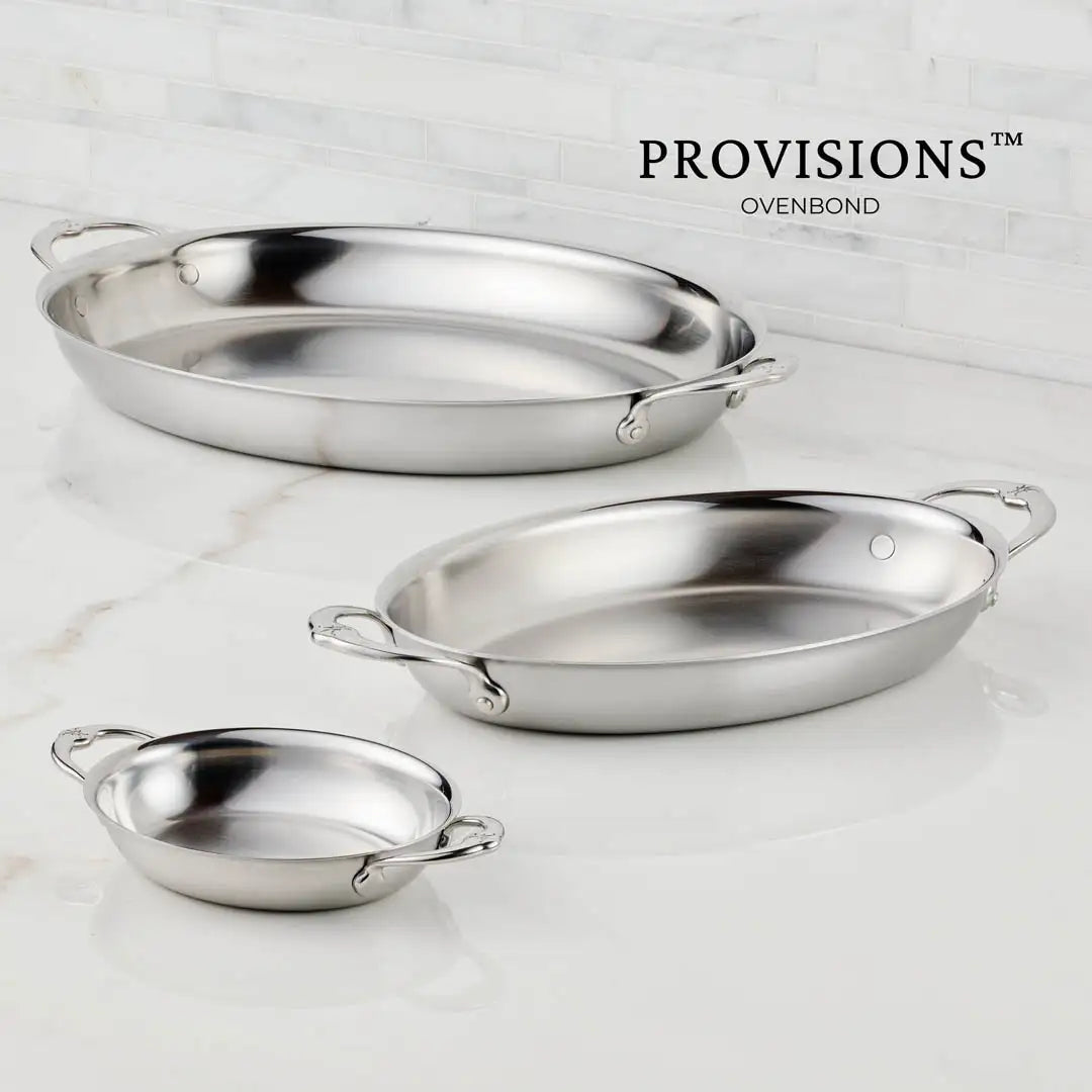 The Hestan Provisions 17.5-Inch x 14-Inch Clad Oval Roaster with handles is displayed on a white marble countertop, showcasing its elegance and "PROVISIONS OVENBOND" label in the background.