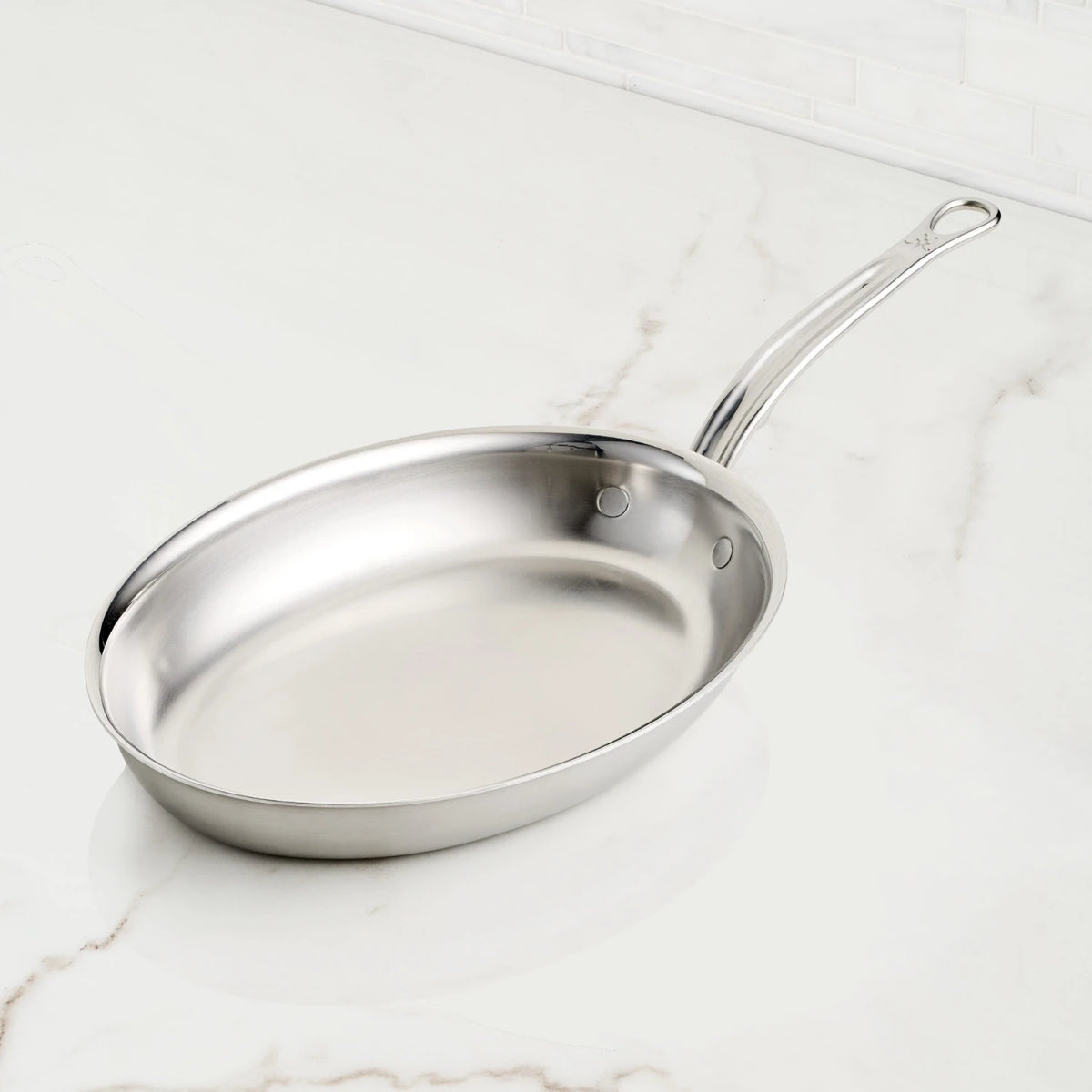 The Oval Fish Pan by Hestan Provisions, known for even heat distribution, features a polished finish and riveted handle. Its positioned slightly angled to the right on a white marble countertop.