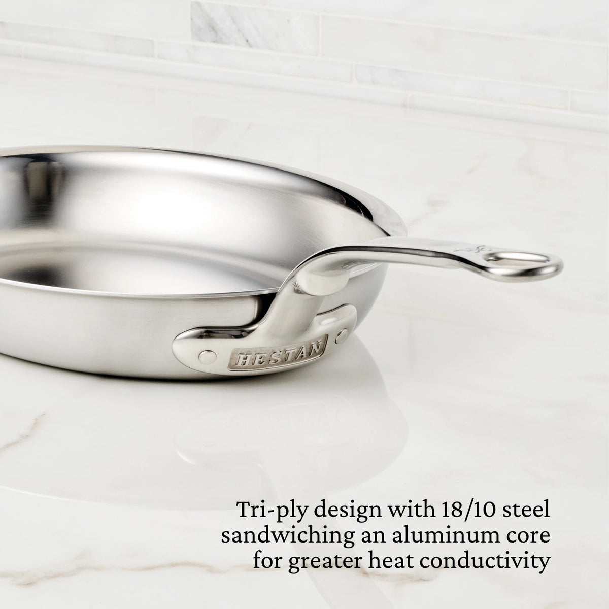 The Hestan Provisions Oval Fish Pan features a silver finish and tri-ply design, with an engraved handle. Corner text highlights its 18/10 stainless steel construction and aluminum core for even heat distribution, resting on a white marble surface.