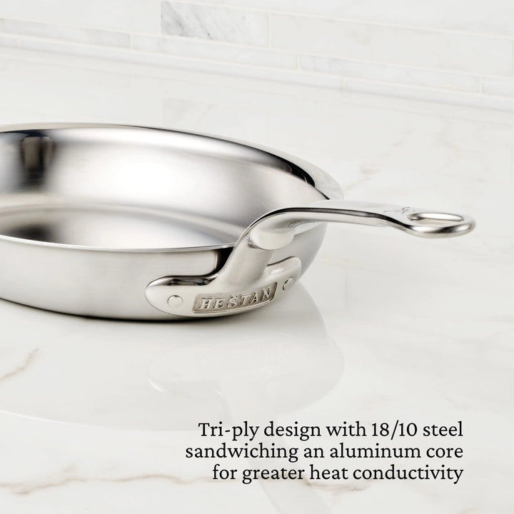 A Thomas Keller Insignia Commercial Clad Stainless Steel 29-Piece Cookware Set sits on a marble countertop, featuring tri-ply 18/10 steel with an aluminum core for excellent heat conductivity and consistent cooking results.