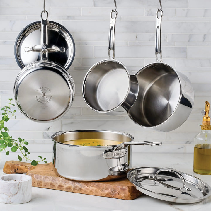Hestan Provisions Stainless Steel Lids are shown in a kitchen with three pieces hanging on hooks against a tiled backsplash and another pot with lid on a wooden board. A plant and olive oil bottle add to the stylish display.