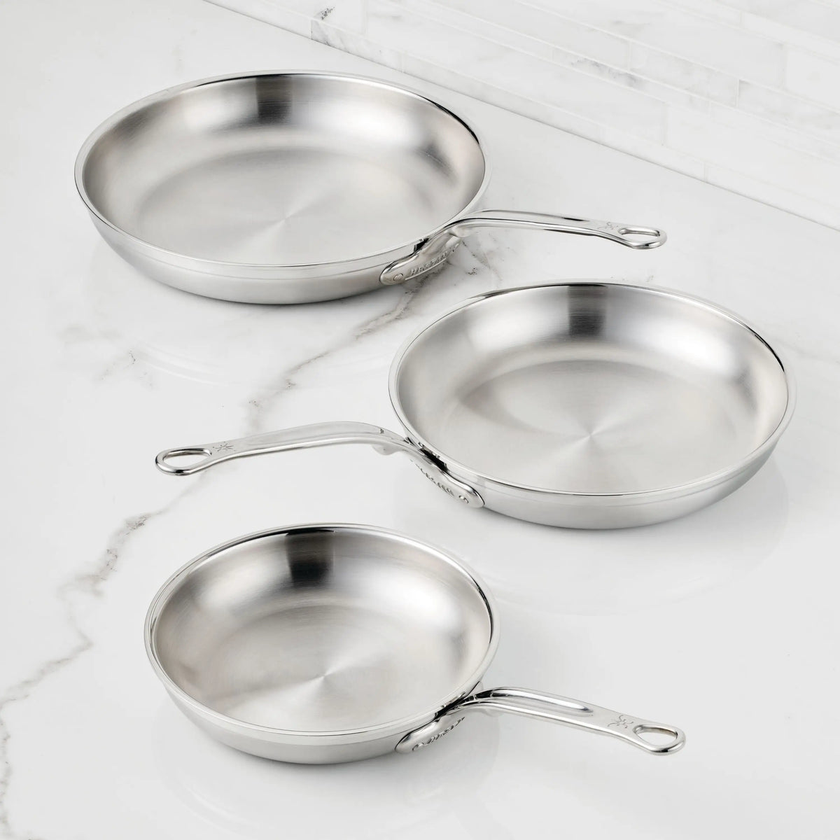 Three ProBond® Forged™ Professional Clad Stainless Steel Skillets, handcrafted in Italy with long metal handles, are arranged on a white marble countertop against a white tiled backsplash.