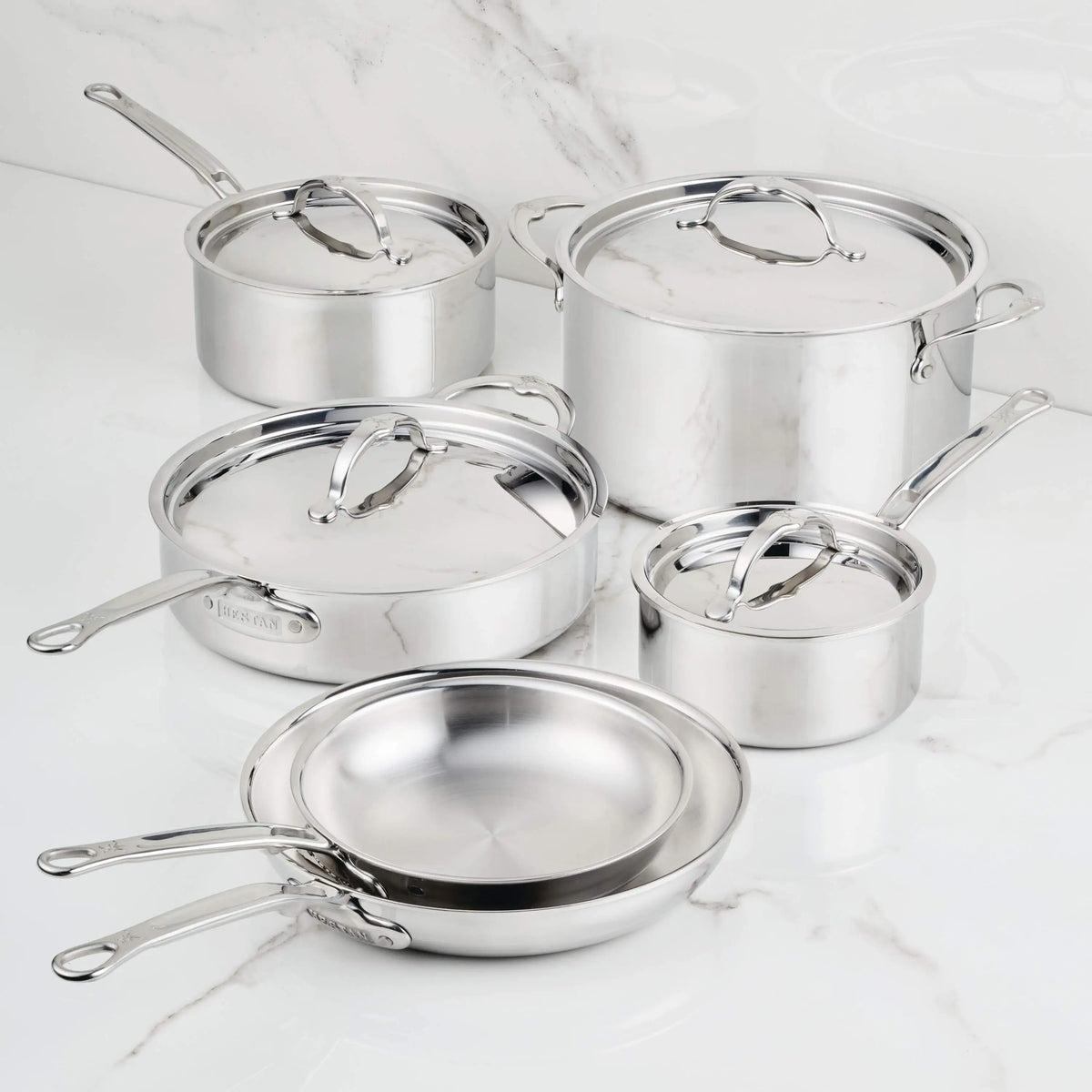 The ProBond® Luxe™ Polished Clad Stainless Steel Ultimate Set, 10-Piece, features saucepans, a frying pan, and a stock pot with lids—each boasting polished surfaces and metal handles, arranged on a white marble countertop.