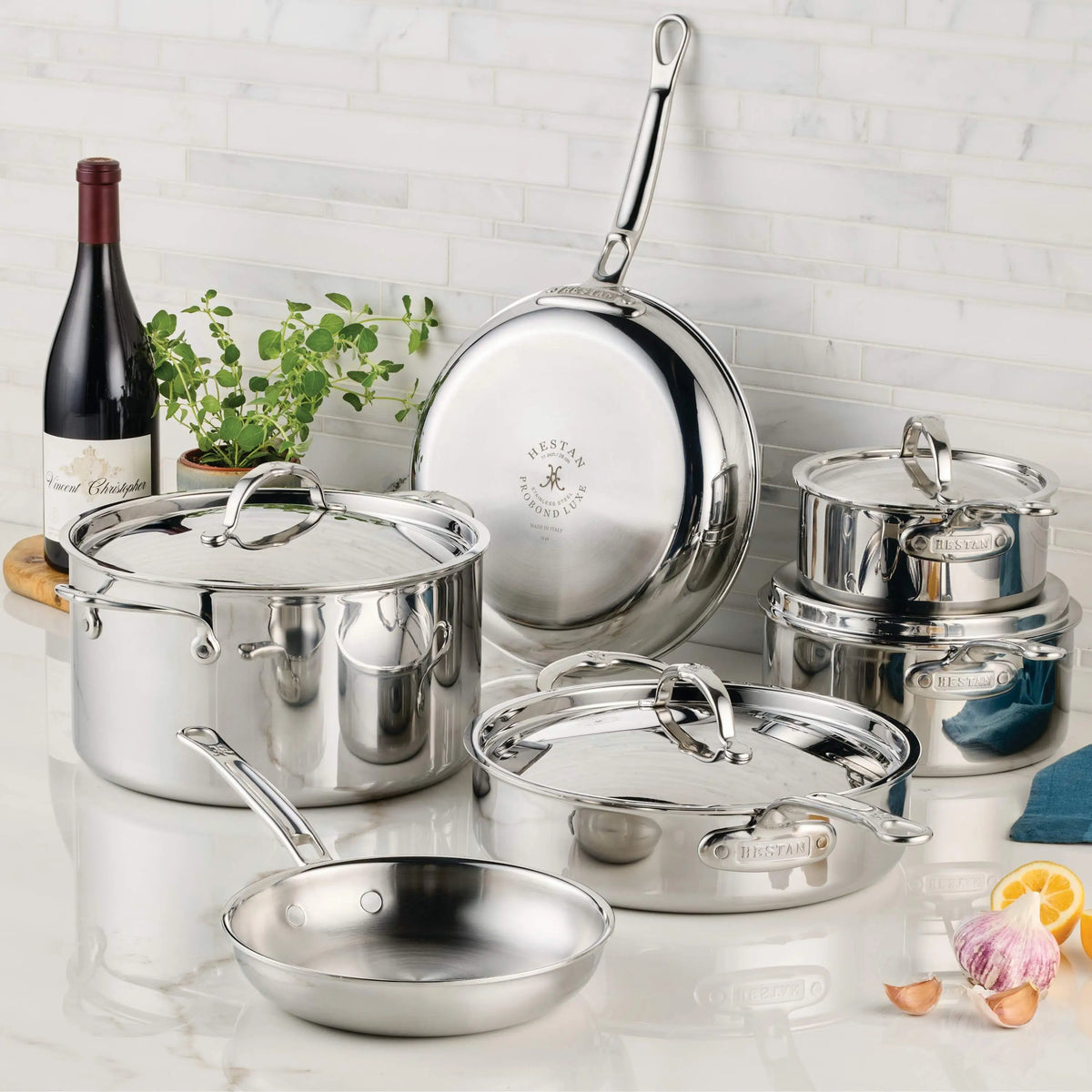 A ProBond® Luxe™ Polished Clad Stainless Steel Ultimate Set, 10-Piece, with lids is displayed on a kitchen counter alongside a bottle of red wine, a potted plant, garlic, shallot, and a blue towel against a white tile wall.