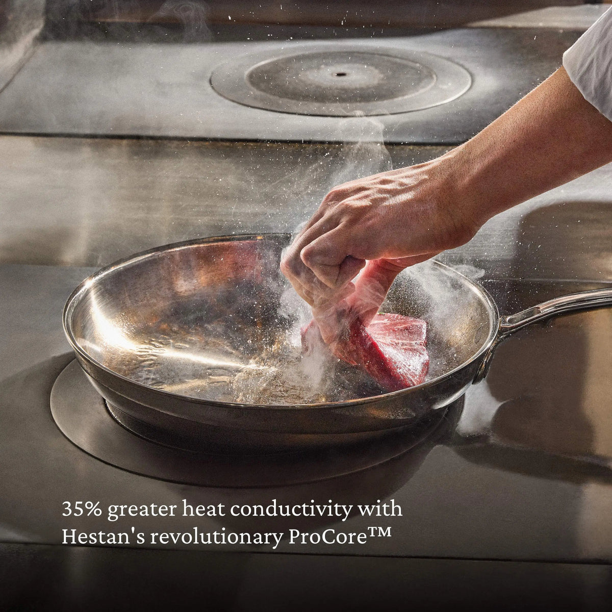 A hand adds raw meat to a stainless steel pan on a stove, sending up steam. Text: ProBond® Luxe™ Polished Clad Stainless Steel Ultimate Set heats 35% better with revolutionary ProCore™ technology.