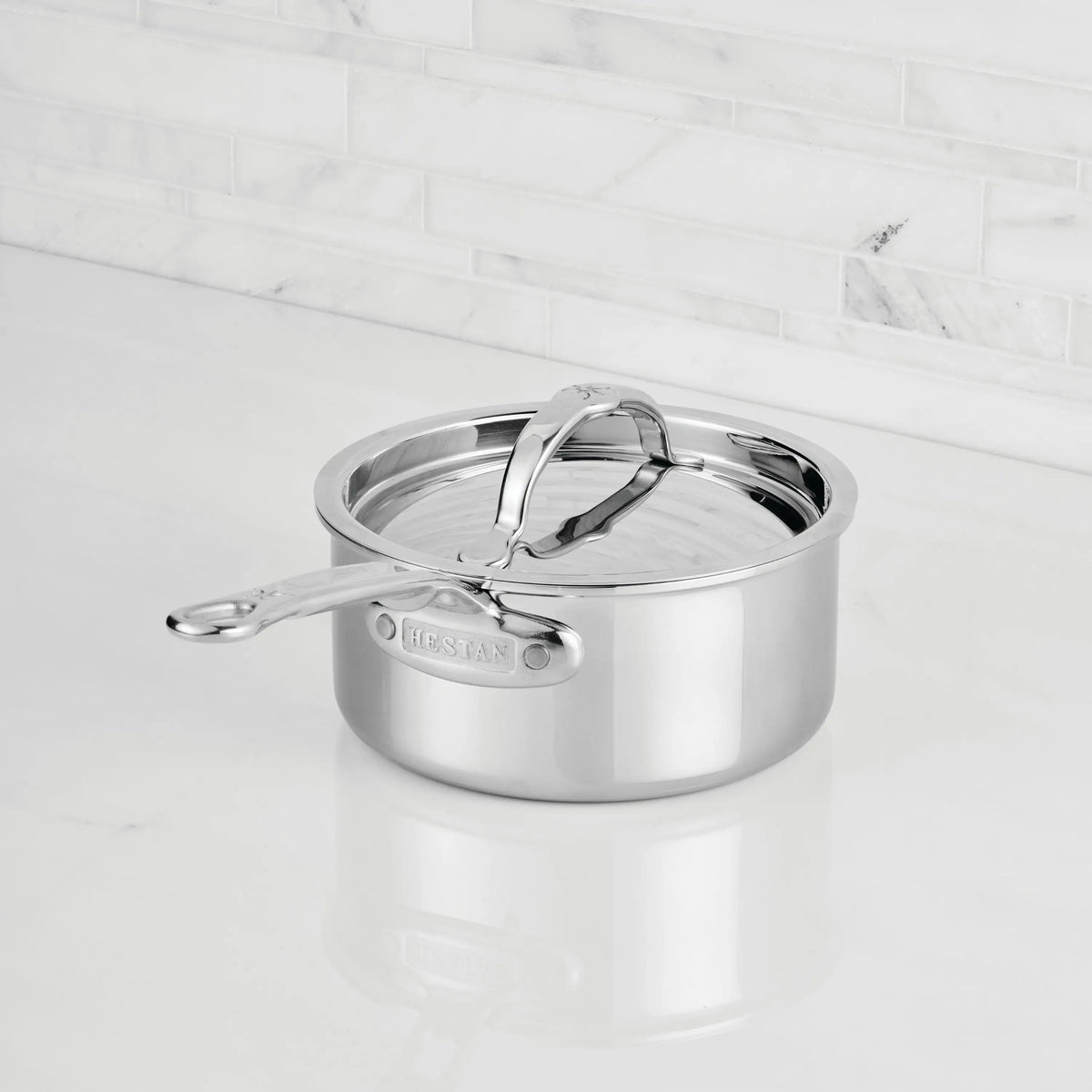A ProBond® Luxe™ Polished Clad Stainless Steel Saucepan with lid sits on a white marble kitchen countertop in front of a white tiled backsplash.