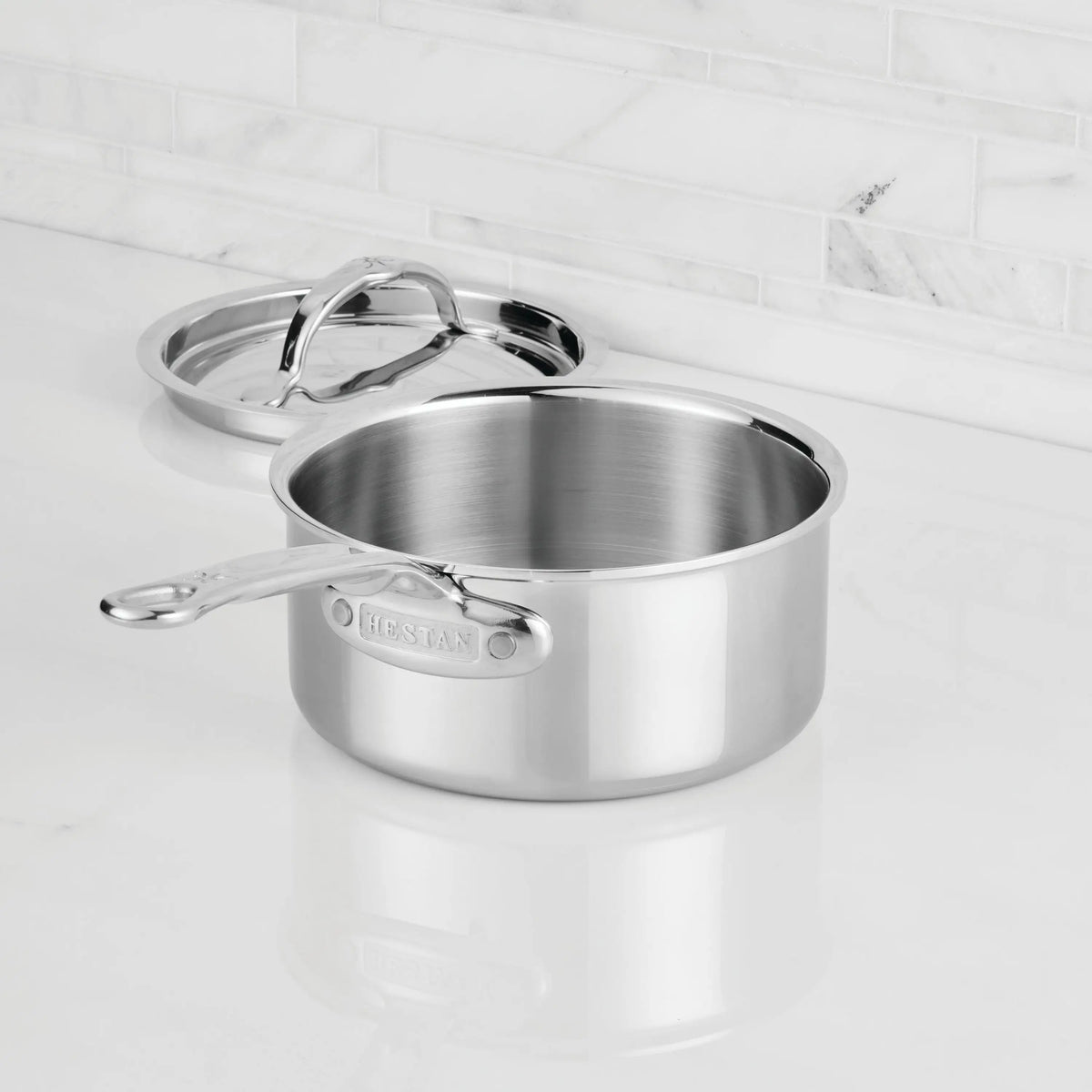 A ProBond® Luxe™ Polished Clad Stainless Steel Saucepan with lid sits on a white countertop in front of a white tiled backsplash. The matching lid rests behind the saucepan, showcasing this premium cookware.