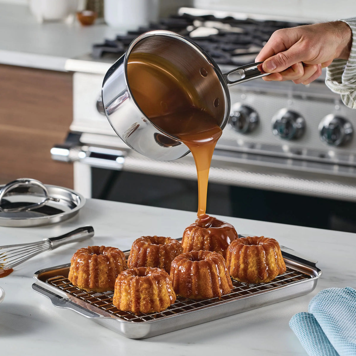 A person pours caramel sauce from a ProBond® Luxe™ Polished Clad Stainless Steel Saucepan with Lid over six bundt cakes on a cooling rack and baking sheet in the kitchen, with stove and utensils visible in the background.